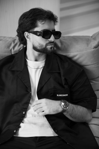 Een man in donkere kleding en zonnebril, zittend op een bank.